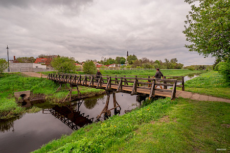 Drewniany most nad niewielką rzeką. Na moście kobieta jedzie rowerem, w przeciwnym kierunku idzie mężczyzna z psem