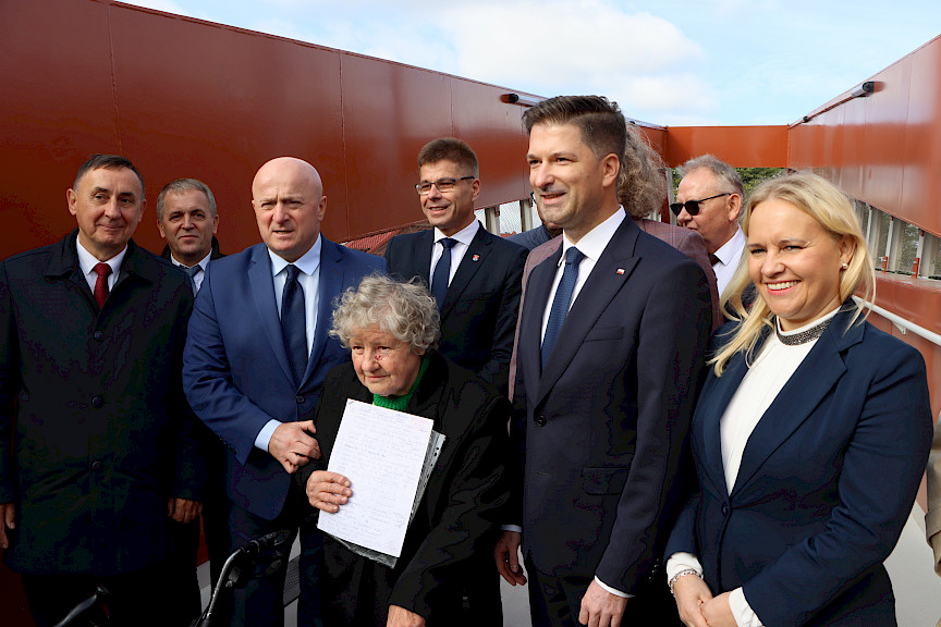 Grupa ludzi w różnym wieku stoi na świeżym powietrzu. Siedmiu oficjalnie ubranych mężczyzn, z prawej strony stoi kobieta. W środku starsza kobieta trzymająca przed sobą dokument.