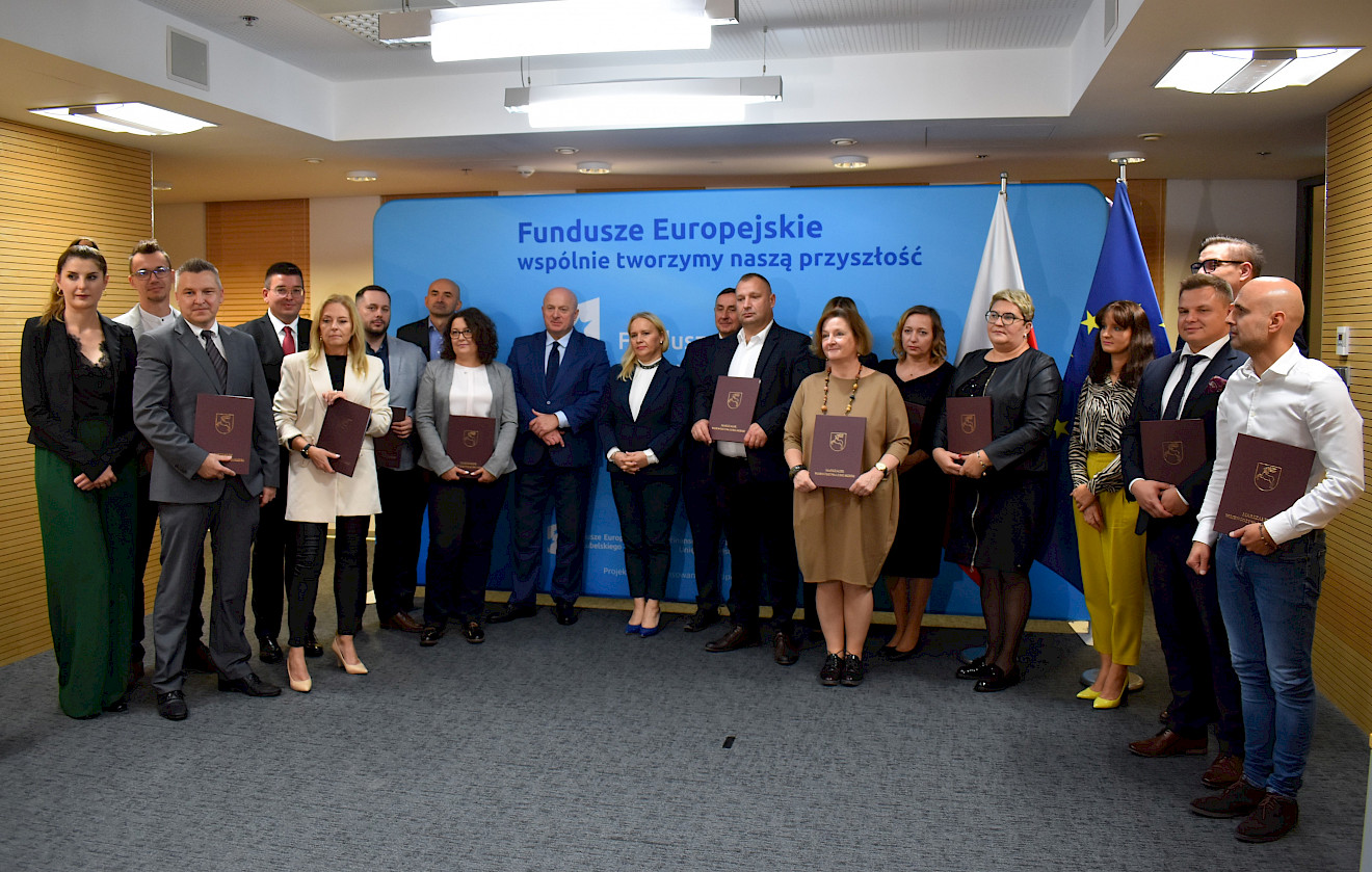 20 osób w różnym wieku obojga płci pozuje do fotografii na tle ścianki z napisem Fundusze Europejskie wspólnie tworzymy naszą przyszłość. Większość spośród nich trzyma brązowe teczki na dokumenty