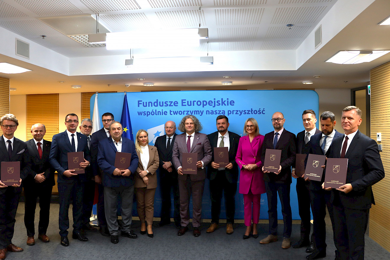 Na ściance promocyjnej Funduszy Europejskich stoi dwunastu mężczyzn - Beneficjentów Funduszy Europejskich trzymając w rękach wręczone dzisiaj umowy. Na zdjęciu także marszałek Jarosław Stawiarski, dyrektor DZ PR Anna Brzyska oraz radna Anna Baluch