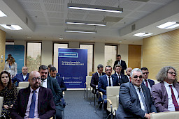 Na widowni przed rozpoczęciem konferencji siedzą na krzesłach Beneficjenci, którzy później odebrali umowy na realizację inwestycji unijnych. W tle stoi niebieski rollup Fundusze Europejskie dla Lubelskiego 2021-2027.