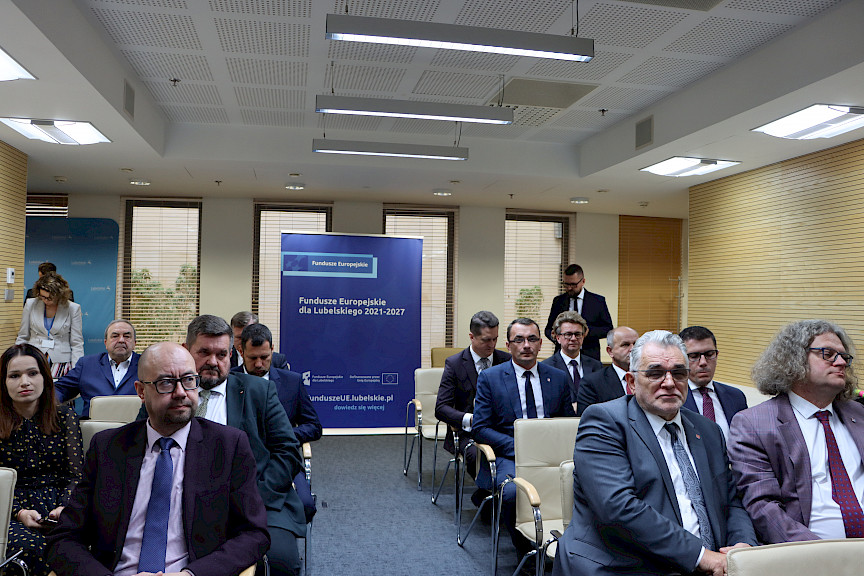 Na widowni przed rozpoczęciem konferencji siedzą na krzesłach Beneficjenci, którzy później odebrali umowy na realizację inwestycji unijnych. W tle stoi niebieski rollup Fundusze Europejskie dla Lubelskiego 2021-2027.