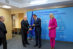Marszałek Jarosław Stawiarki oraz radna Anna Baluch wręczają na tle ścianki Fundusze Europejskie wspólnie tworzymy naszą przyszłość, umowę Beneficjentowi na realizację unijnej inwestycji. Marszałek z Beneficjentem ściskają sobie dłoń na tle flagi Polski i UE.
