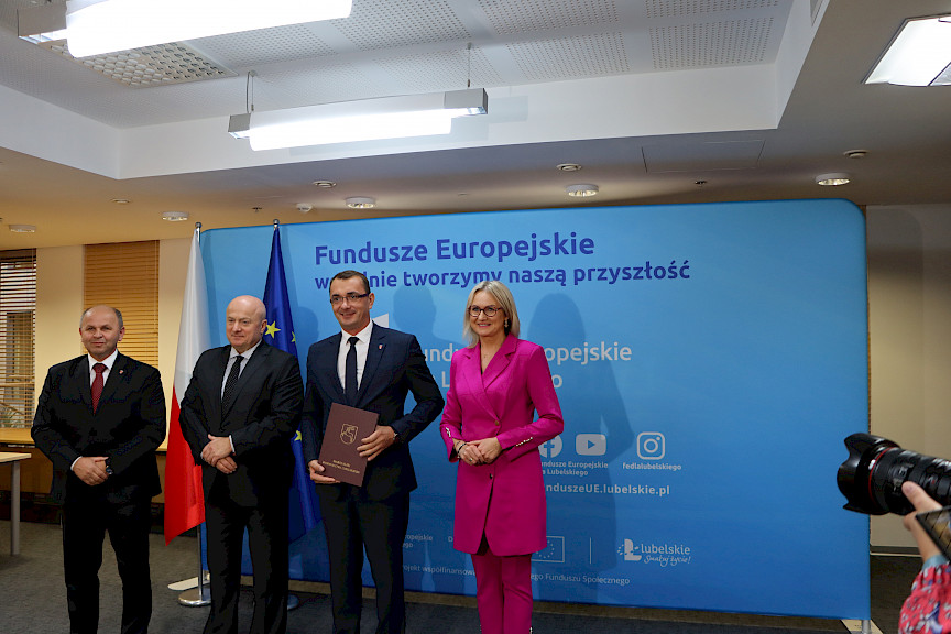 Pamiątkowe zdjęcie marszałka Jarosława Stawiarskiego oraz radnej Anny Baluch w towarzystwie Beneficjenta, któremu wcześniej wręczyli umowę na realizację inwestycji. Wszyscy stoją na tle ścianki ścianki Fundusze Europejskie wspólnie tworzymy naszą przyszłość oraz na tle flagi Polski i UE.