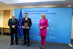 Pamiątkowe zdjęcie marszałka Jarosława Stawiarskiego oraz radnej Anny Baluch w towarzystwie Beneficjenta, któremu wcześniej wręczyli umowę na realizację inwestycji. Wszyscy stoją na tle ścianki ścianki Fundusze Europejskie wspólnie tworzymy naszą przyszłość oraz na tle flagi Polski i UE.