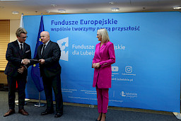 Pamiątkowe zdjęcie marszałka Jarosława Stawiarskiego oraz radnej Anny Baluch w towarzystwie Beneficjenta. Wszyscy stoją na tle ścianki ścianki Fundusze Europejskie wspólnie tworzymy naszą przyszłość. Marszałek z Beneficjentem ściskają sobie dłoń na tle flagi Polski i UE. Marszałek wręcza Beneficjentowi umowę.