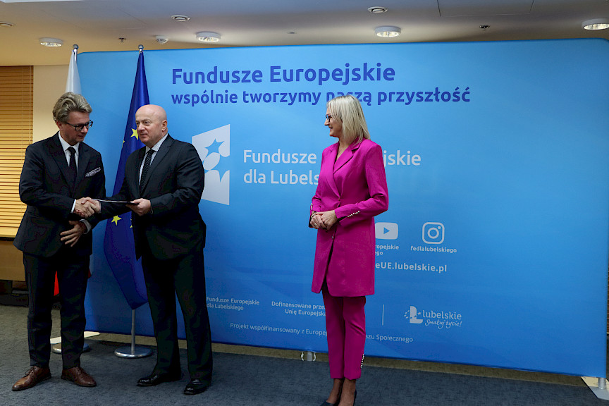 Pamiątkowe zdjęcie marszałka Jarosława Stawiarskiego oraz radnej Anny Baluch w towarzystwie Beneficjenta. Wszyscy stoją na tle ścianki ścianki Fundusze Europejskie wspólnie tworzymy naszą przyszłość. Marszałek z Beneficjentem ściskają sobie dłoń na tle flagi Polski i UE. Marszałek wręcza Beneficjentowi umowę.