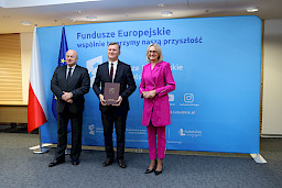 Pamiątkowe zdjęcie marszałka Jarosława Stawiarskiego oraz radnej Anny Baluch w towarzystwie Beneficjenta. Wszyscy stoją na tle ścianki ścianki Fundusze Europejskie wspólnie tworzymy naszą przyszłość oraz na tle flagi Polski i UE. Beneficjent w ręku w teczce trzyma przed sobą otrzymaną umowę.