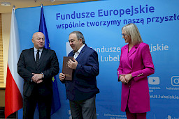 Pamiątkowe zdjęcie marszałka Jarosława Stawiarskiego oraz radnej Anny Baluch w towarzystwie Beneficjenta. Wszyscy stoją na tle ścianki ścianki Fundusze Europejskie wspólnie tworzymy naszą przyszłość oraz na tle flagi Polski i UE. Beneficjent w ręku w teczce trzyma przed sobą otrzymaną umowę.