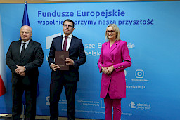 Pamiątkowe zdjęcie marszałka Jarosława Stawiarskiego oraz radnej Anny Baluch w towarzystwie Beneficjenta. Wszyscy stoją na tle ścianki ścianki Fundusze Europejskie wspólnie tworzymy naszą przyszłość oraz na tle flagi Polski i UE. Beneficjent w ręku w teczce trzyma przed sobą otrzymaną umowę.