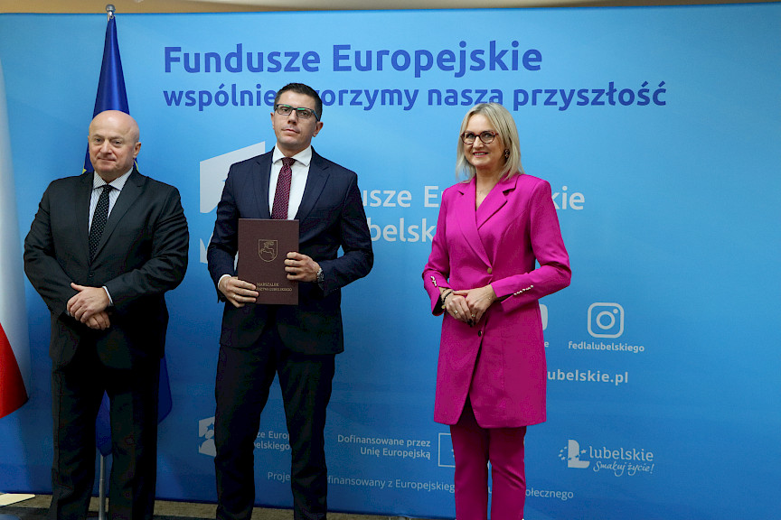 Pamiątkowe zdjęcie marszałka Jarosława Stawiarskiego oraz radnej Anny Baluch w towarzystwie Beneficjenta. Wszyscy stoją na tle ścianki ścianki Fundusze Europejskie wspólnie tworzymy naszą przyszłość oraz na tle flagi Polski i UE. Beneficjent w ręku w teczce trzyma przed sobą otrzymaną umowę.