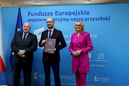 Pamiątkowe zdjęcie marszałka Jarosława Stawiarskiego oraz radnej Anny Baluch w towarzystwie Beneficjenta. Wszyscy stoją na tle ścianki ścianki Fundusze Europejskie wspólnie tworzymy naszą przyszłość oraz na tle flagi Polski i UE. Beneficjent w ręku w teczce trzyma przed sobą otrzymaną umowę.