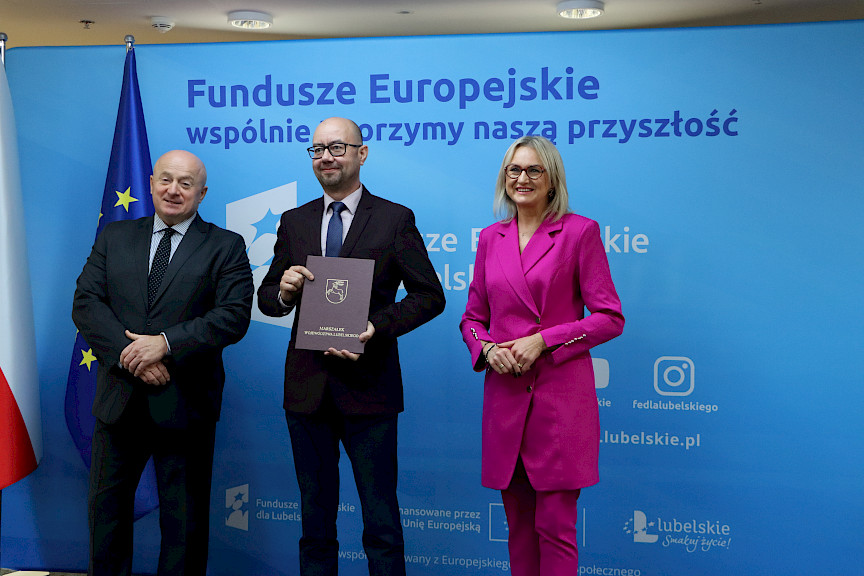 Pamiątkowe zdjęcie marszałka Jarosława Stawiarskiego oraz radnej Anny Baluch w towarzystwie Beneficjenta. Wszyscy stoją na tle ścianki ścianki Fundusze Europejskie wspólnie tworzymy naszą przyszłość oraz na tle flagi Polski i UE. Beneficjent w ręku w teczce trzyma przed sobą otrzymaną umowę.