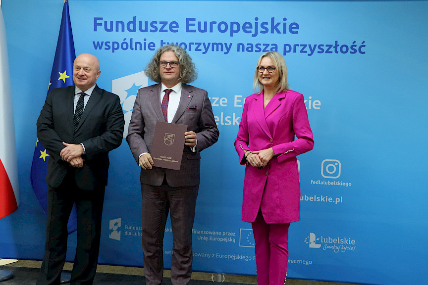 Pamiątkowe zdjęcie marszałka Jarosława Stawiarskiego oraz radnej Anny Baluch w towarzystwie Beneficjenta. Wszyscy stoją na tle ścianki ścianki Fundusze Europejskie wspólnie tworzymy naszą przyszłość oraz na tle flagi Polski i UE. Beneficjent w ręku w teczce trzyma przed sobą otrzymaną umowę.