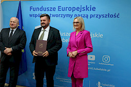 Pamiątkowe zdjęcie marszałka Jarosława Stawiarskiego oraz radnej Anny Baluch w towarzystwie Beneficjenta. Wszyscy stoją na tle ścianki ścianki Fundusze Europejskie wspólnie tworzymy naszą przyszłość oraz na tle flagi Polski i UE. Beneficjent w ręku w teczce trzyma przed sobą otrzymaną umowę.