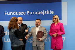 Na tle niebieskiej ścianki Fundusze Europejskie dla Lubelskiego 2021-2027 stoi marszałek Jarosław Stawiarski, radna Anna Baluch i Beneficjent z odebraną przed chwilą umową, którzy udziela wywiadu lokalnym mediom