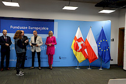 Na tle niebieskiej ścianki Fundusze Europejskie dla Lubelskiego 2021-2027 stoi marszałek Jarosław Stawiarski, radna Anna Baluch i Beneficjent z odebraną przed chwilą umową, którzy udziela wywiadu lokalnym mediom