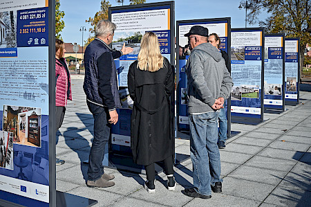 Wystawa prezentująca efekty wdrażania Funduszy Europejskich w województwie lubelskim na rynku w Janowie Lubelskim oraz osoby ją oglądające.