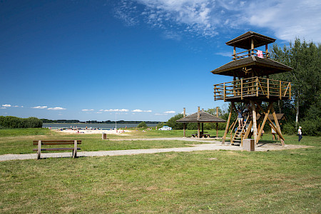 Na pierwszym planie drewniana wieża widokowa, z której schodzą ludzie. W głębi plaża, na której wypoczywają ludzie. Za nią rozciąga się jezioro