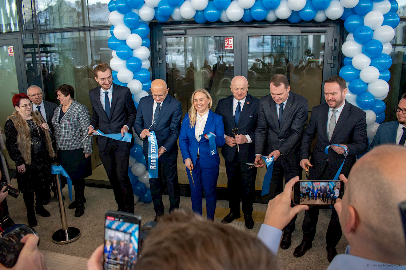 Otwarcie Dworca Lublin.  Grupa oficjalnie ubranych osób z fragmentami przeciętej wstęgi. W tle przeszklony budynek, do którego drzwi otoczone zostały konstrukcją z biało niebieskich baloników