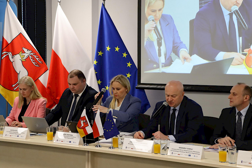 Trzech mężczyzn i dwie kobiety siedzą przy stole prezydialnym, jeden z mężczyzn przemawia do mikrofonu, w tle ekran z tytułem konferencji, po bokach flagi województwa lubelskiego, Polski i UE