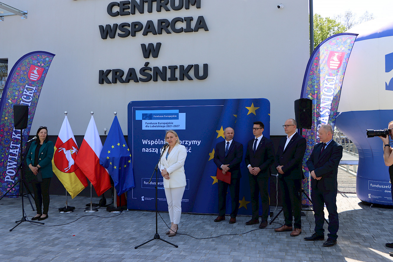 Sześć osób stojących na niebieskiej ściance promocyjnej. Jedna kobieta przemawia do mikrofonu