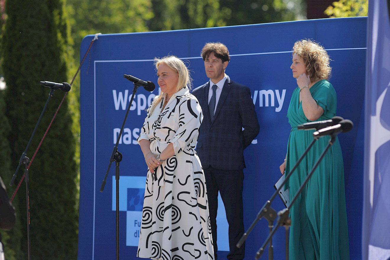 Trzy osoby stoją na scenie z niebieskim tłem. Kobieta w biało-czarnej wzorzystej sukience przemawia do mikrofonu. Obok niej stoją mężczyzna w niebieskim garniturze i kobieta w zielonej sukience. Widoczne są dwa dodatkowe mikrofony, w tle drzewa