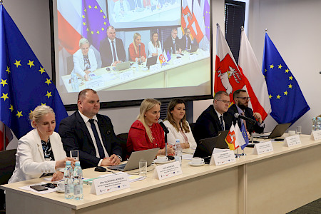 Sześć oficjalnie ubranych osób (trzy kobiety) siedzi za stołem prezydialnym. Powyżej ekran wyświetlający ten sam obraz