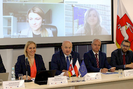 Obraz pokazuje panel dyskusyjny z siedzącymi osobami. Na środku starszy mężczyzna przemawia do mikrofonu, obok niego kobieta i inni mężczyźni. Na ekranie widać dwie młode kobiety, sugerując wideokonferencję. Widoczne flagi i logotypy instytucjonalne