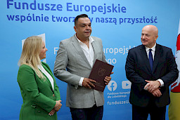 Marszałek Województwa Lubelskiego, Pan Jarosław Stawiarski wraz z Panią Anną Brzyską, Dyrektor Departamentu Zarządzania Programami Regionalnymi z Beneficjentem, mężczyzną w garniturze pozują po wręczeniu umowy na tle ścianki promocyjnej Fundusze Europejskie dla Lubelskiego