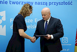Marszałek Województwa Lubelskiego, Pan Jarosław Stawiarski wręcza umowę i ściska dłoń Beneficjentki na tle ścianki promocyjnej  Fundusze Europejskie dla Lubelskiego
