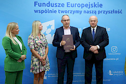 Marszałek Województwa Lubelskiego, Pan Jarosław Stawiarski wraz z Panią Anną Brzyską, Dyrektor Zarządzania Programami Regionalnymi pozują z Beneficjentami, kobietą i mężczyzną na tle ścianki promocyjnej  Fundusze Europejskie dla Lubelskiego.