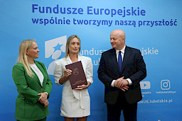 Marszałek Województwa Lubelskiego, Pan Jarosław Stawiarski wraz z Panią Anną Brzyską, Dyrektor Departamentu Zarządzania Programami Regionalnymi pozują  z Beneficjentką na tle ścianki promocyjnej  Fundusze Europejskie dla Lubelskiego.