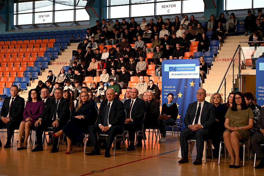 Uroczyste wręczenie umów z Działania 10.4 w Radzyniu Podlaskim w dniu 21.10.2024 r. Wszyscy zgromadzeni beneficjenci siedzą na krzesłach na sali gimnastycznej. za nimi siedzą zgromadzeniu uczniowie szkoły.