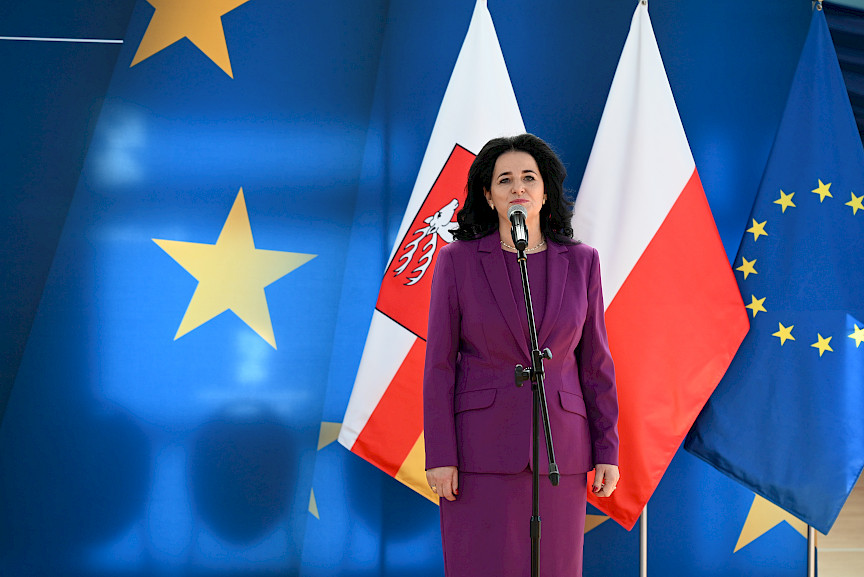 Uroczyste wręczenie umów z Działania 10.4 w Radzyniu Podlaskim w dniu 21.10.2024 r. Dyrektor szkoły, w której wręczano umowy na tle ścianki funduszy europejskich przemawia do zgromadzonych gości.