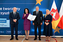 Uroczyste wręczenie umów z Działania 10.4 w Radzyniu Podlaskim w dniu 21.10.2024 r. Marszałek, dyrektor szkoły, starosta radzyński i dyrektor Departamentu Zarządzania Programami Regionalnymina stoją na tle ścianki funduszy europejskich przemawia do zgromadzonych gości.