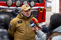 Uroczyste przekazanie sprzętu Straży Pożarnej w Kraśniku, strażak udziela wywiadu TVP i Dziennikowi Wschodniemu, w tle przekazany wóz strażacki.