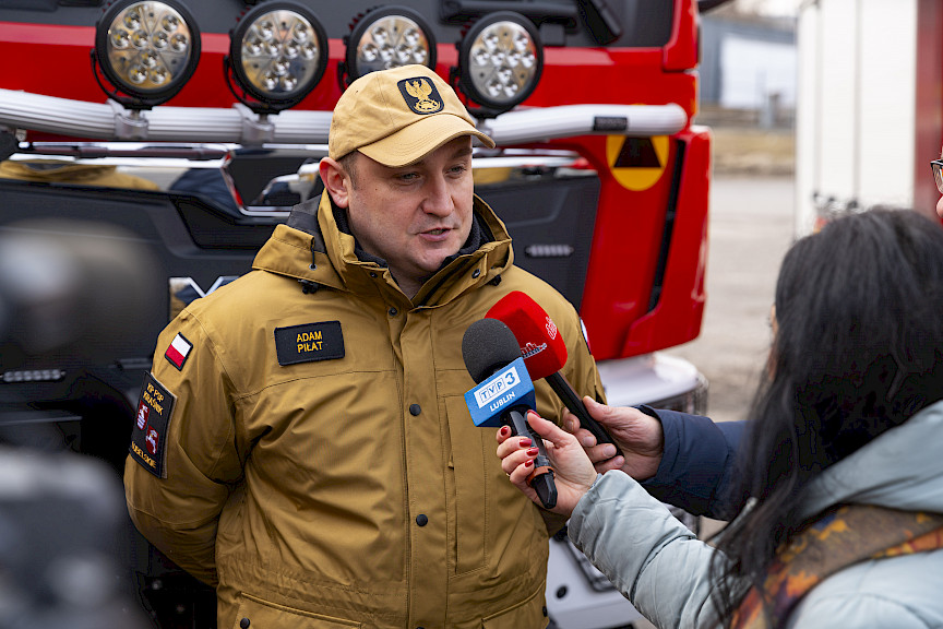 Uroczyste przekazanie sprzętu Straży Pożarnej w Kraśniku, strażak udziela wywiadu TVP i Dziennikowi Wschodniemu, w tle przekazany wóz strażacki.