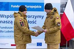 Uroczyste przekazanie sprzętu Straży Pożarnej w Kraśniku, komendant wręcza umowę strażakowi, w tle ścianka Funduszy Europejskich, po prawej flaga Polski.