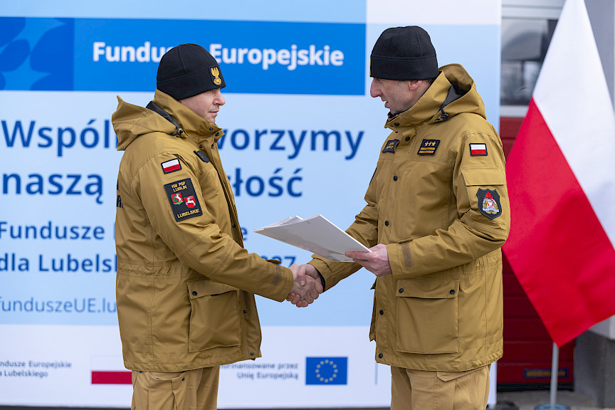 Uroczyste przekazanie sprzętu Straży Pożarnej w Kraśniku, komendant wręcza umowę strażakowi, w tle ścianka Funduszy Europejskich, po prawej flaga Polski.