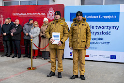 Uroczyste przekazanie sprzętu Straży Pożarnej w Kraśniku, strażacy pozują z wręczonymi umowami, w tle ścianka Funduszy Europejskich.