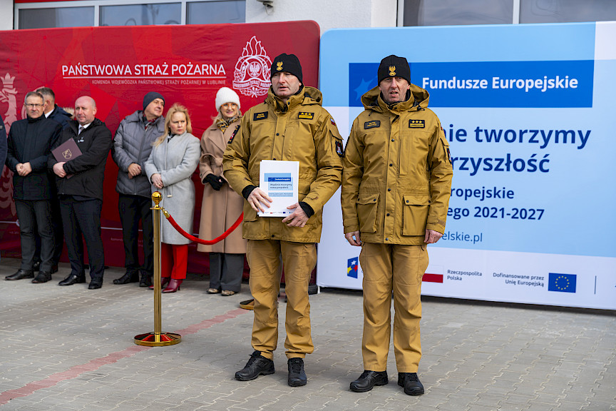 Uroczyste przekazanie sprzętu Straży Pożarnej w Kraśniku, strażacy pozują z wręczonymi umowami, w tle ścianka Funduszy Europejskich.