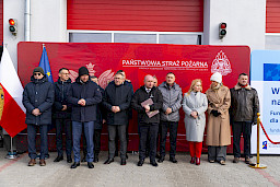 Uroczyste przekazanie sprzętu Straży Pożarnej w Kraśniku, zaproszeni goście stoją na tle ścianki Państwowej Straży Pożarnej.