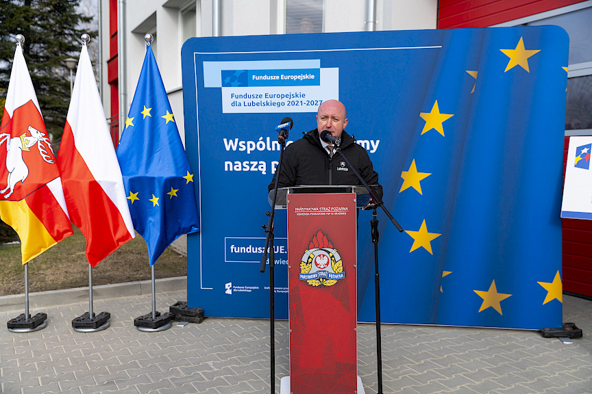 Uroczyste przekazanie sprzętu Straży Pożarnej w Kraśniku, Wicemarszałek Piotr Breś przemawia do strażaków oraz zgromadzonych gości, w tle ścianka Funduszy Europejskich, po lewej flagi Województwa Lubelskiego, Polski, Unii Europejskiej.