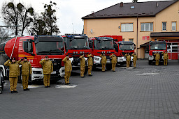 Uroczyste wręczenie sprzętu Straży Pożarnej. Strażacy stojący przed przekazywanymi wozami strażackimi.