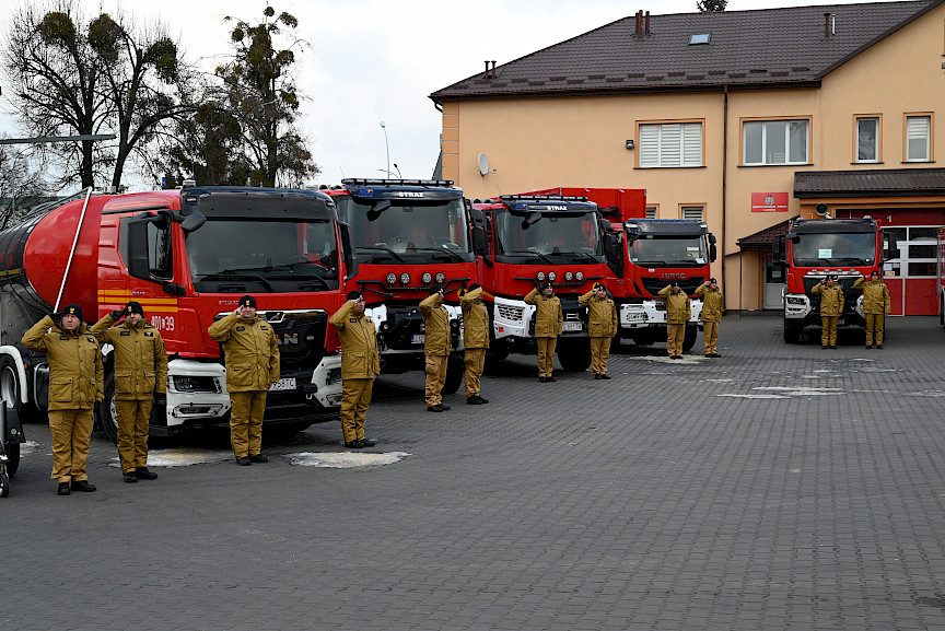 Uroczyste wręczenie sprzętu Straży Pożarnej. Strażacy stojący przed przekazywanymi wozami strażackimi.