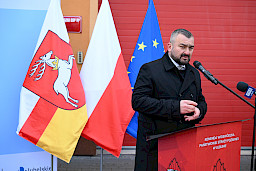 Uroczyste wręczenie sprzętu Straży Pożarnej. Członek Zarządu Województwa Lubelskiego przemawia do zgromadzonych gości. Za plecami flaga Województwa Lubelskiego, Polski oraz Unii Europejskiej.