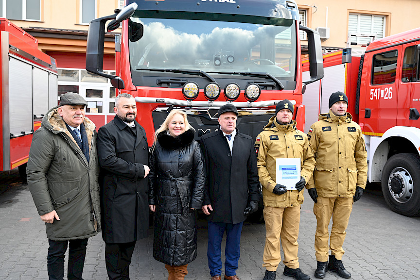 Uroczyste wręczenie sprzętu Straży Pożarnej. Zebrani goście na tle przekazywanego wozu strażackiego.