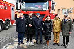 Uroczyste wręczenie sprzętu Straży Pożarnej. Zebrani goście na tle przekazywanego wozu strażackiego.