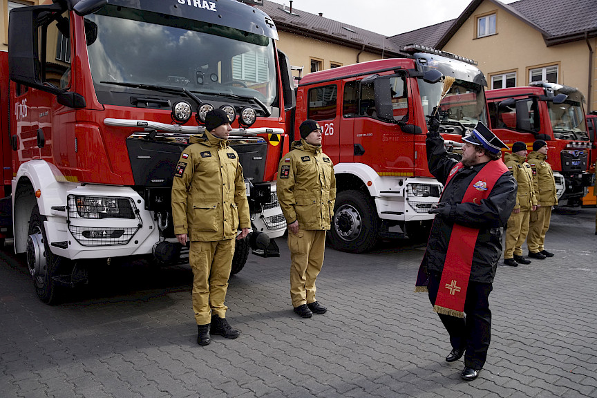Uroczyste wręczenie sprzętu Straży Pożarnej. Strażacy stojący przed przekazywanymi wozami strażackimi, ksiądz poświęca przekazany sprzęt.